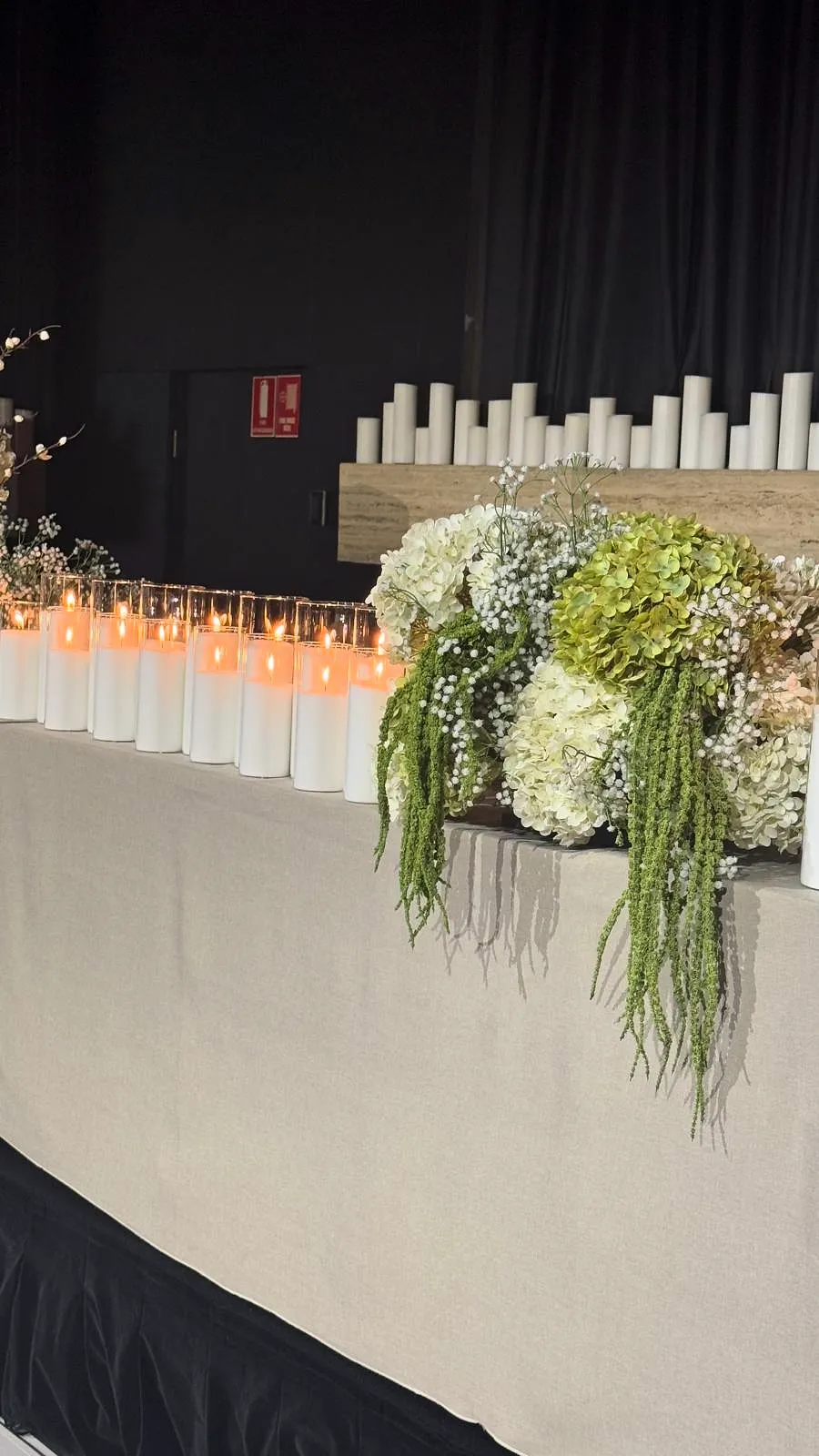 Wedding stage edge with rows of pillar candles, white hydrangeas, gypsophila and trailing green amaranthus