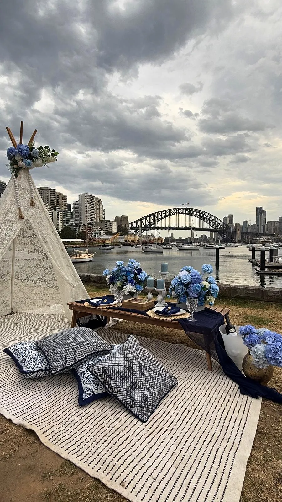 Lace teepee and low table with blue florals on the harbour foreshore under a cloudy sky, with the Harbour Bridge in the distance