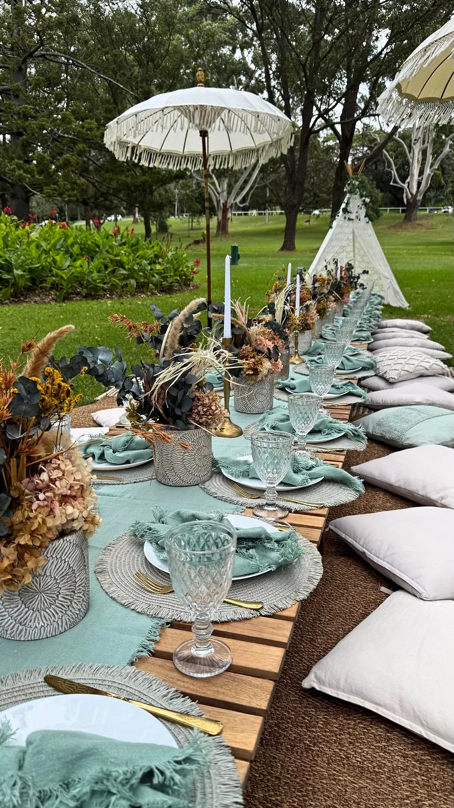 Sage green picnic with dried florals, glassware and fringed umbrellas in a park