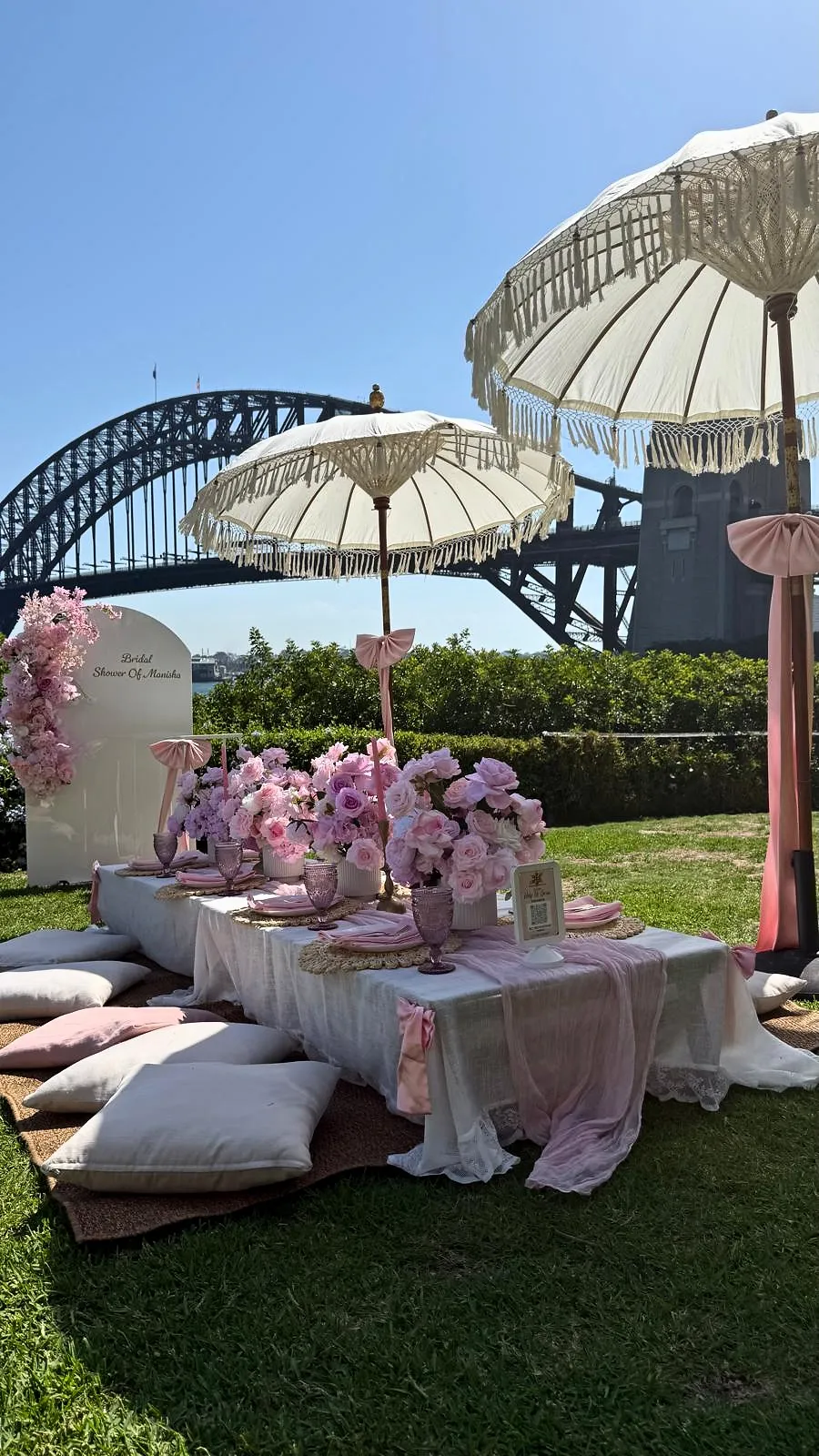 Pink picnic with low tables and cushions under fringed umbrellas, with Sydney Harbour Bridge behind