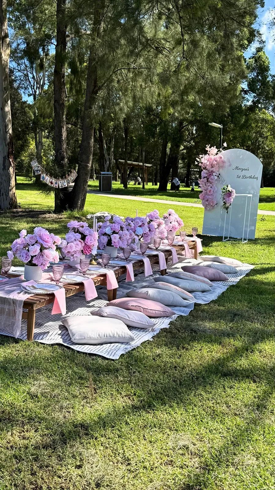 Long lilac and pink picnic table with cushions in a leafy park, with a welcome sign at the end