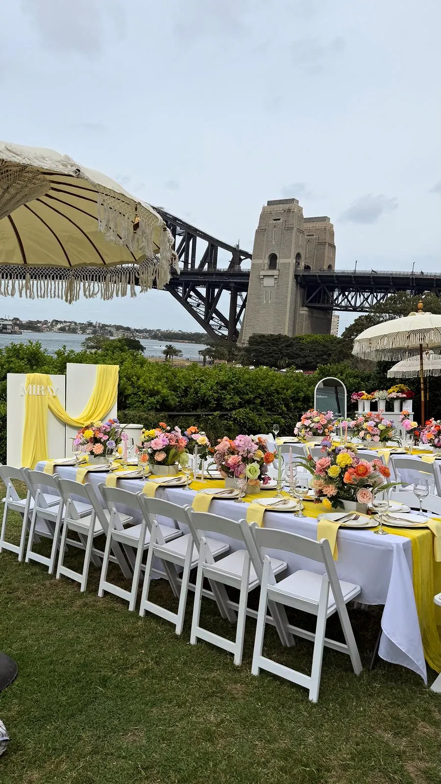Long table with yellow linen and colourful florals under fringed umbrellas, with Sydney Harbour Bridge behind