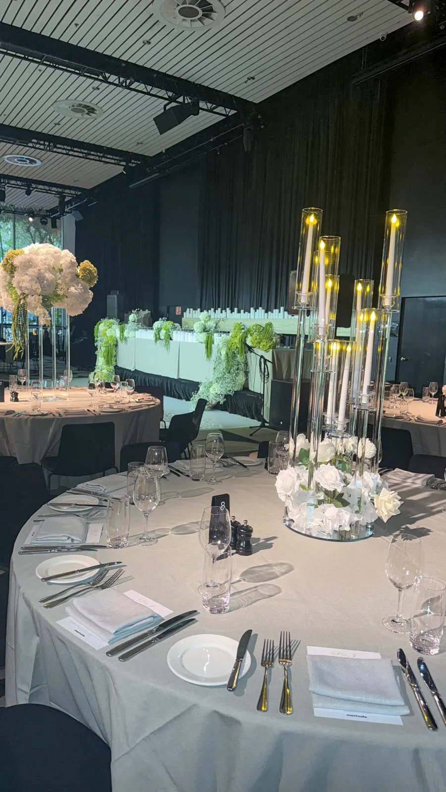 Elegant wedding reception with white table linen, tall glass candleholders, white floral centrepieces and a floral stage against black drapery