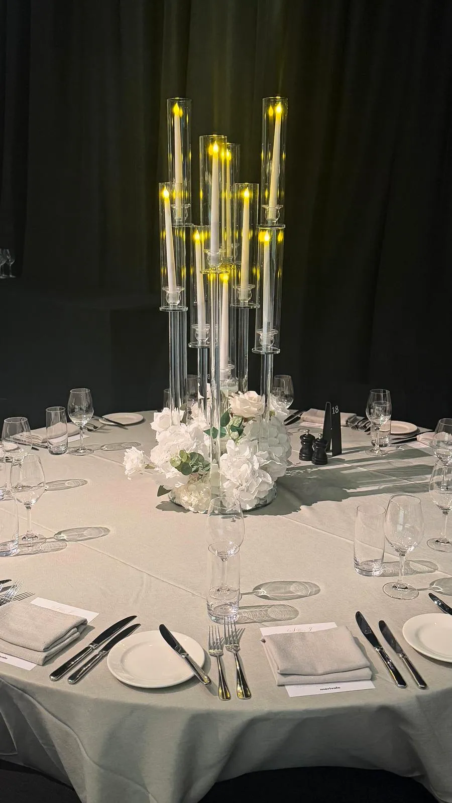 Reception table with a tall glass candelabra rising from white roses and hydrangeas, set against black drapery